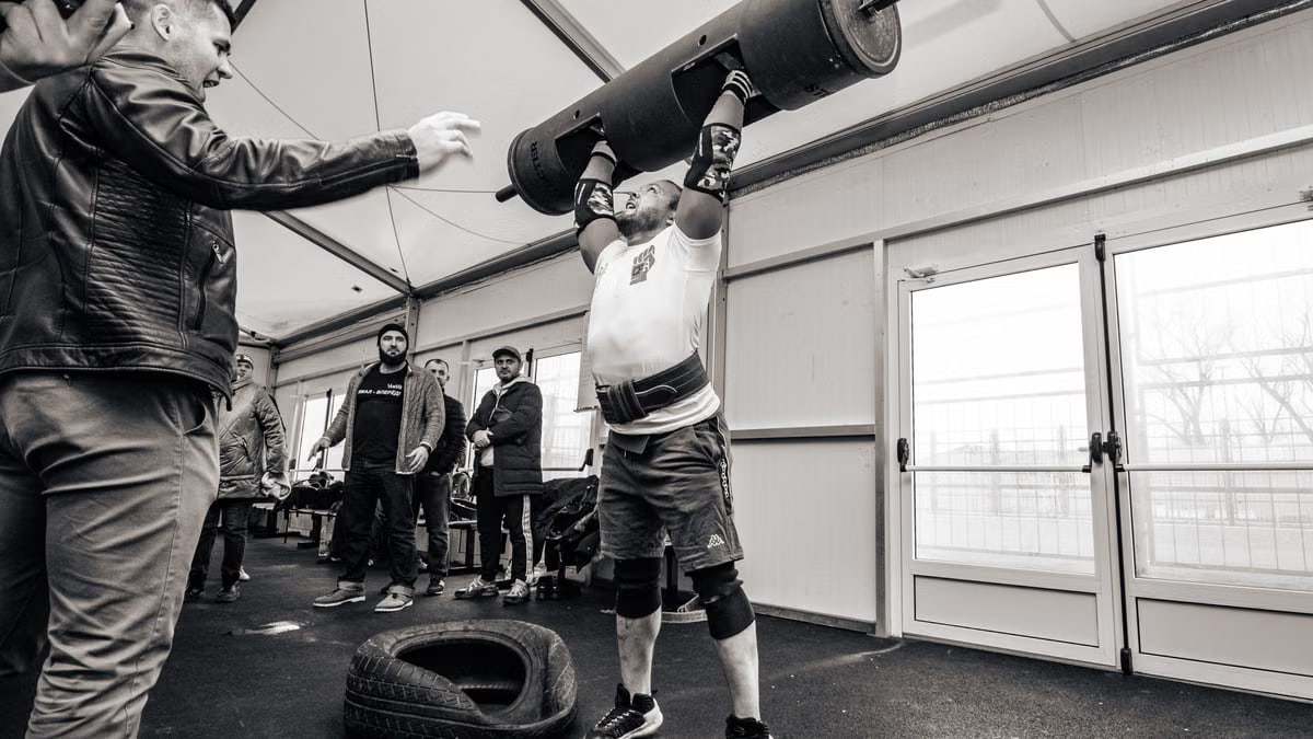A strongman lifts a barbell overhead during an indoor gym event, showcasing strength and determination.